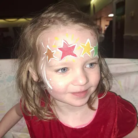 Family handout Beatrix with her forehead painted with stars - gold, red, yellow and green. She has shoulder-length brown hair and is wearing a red top.