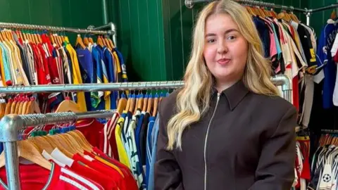 An 18-year-old girl with long blonde hair is looking directly at the camera as she's stood in her store surrounded by hundreds of old football shirts
