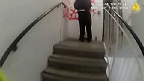 A still image from a police body-worn camera, showing a flight of stairs, and at the top of the stairs, a person wearing black with their back to the camera, and some red graffiti on the white wall.