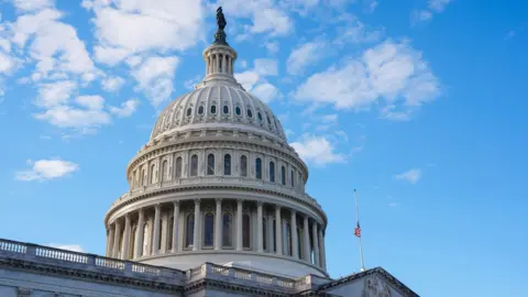 Image shows US Capitol dome against a blue sky with partial cloud cover and the US flag flying in the foreground. Photo taken in Washington DC on Monday.