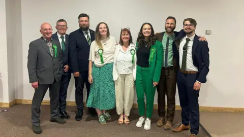Eight people dressed in suits and formal clothes with green rosettes stand together. 