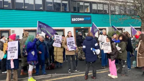 Striking workers stands on a Unison picket line outside Second Step HQ in Bristol. They are holding Unison flags and placards with slogans.