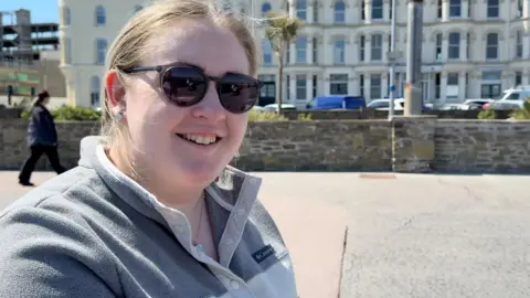 Melanie Harris has light brown hair tied up and has sunglasses on. She is wearing a grey fleece jumper and is smiling. She's standing on a concrete walkway with a stone wall behind her, with terraced buildings and parked cars beyond that, on a sunny day.