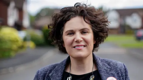 PA Media Claire Hanna, SDLP MP smiles at the camera. She has curly short brown hair and wears a blue blazer with a chunky necklace