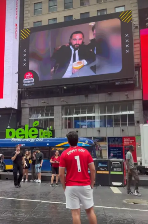 Facebook A man is pictured wearing a red top, looking up at a screen in Times Square in New York which has a picture showing Jimmy Ward 