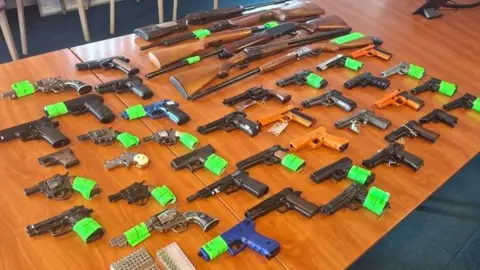 About 45 guns labelled with neon markers laid out on a wooden table.