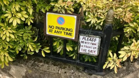 Grace Wood/BBC Home made no parking signs tied to a gate in front of a yellow and green shrub.