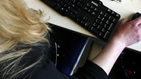 An image taken from above of an unidentified woman with long blonde hair operating a computer mouse at a keyboard