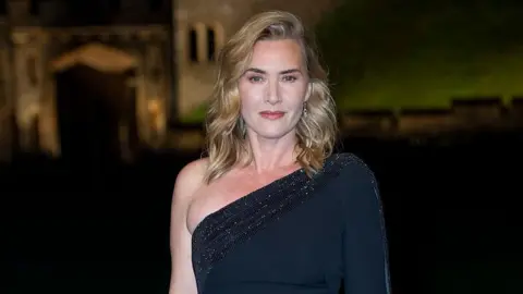 Getty Images Kate smiles while looking at the camera. She is wearing a off the shoulder black sparkly dress and has blonde curly hair.