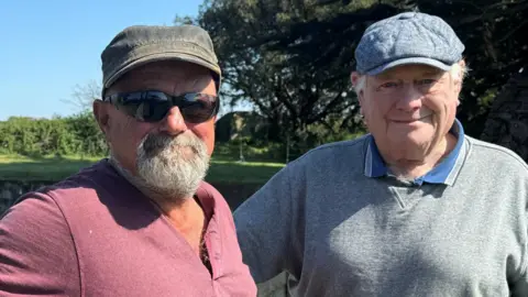 A picture of two men stood smiling at the camera. The man on the left has a hat, a grey beard and black sunglasses. He is wearing a red shirt. The man on the right has a grey hat and a blue and grey shirt.