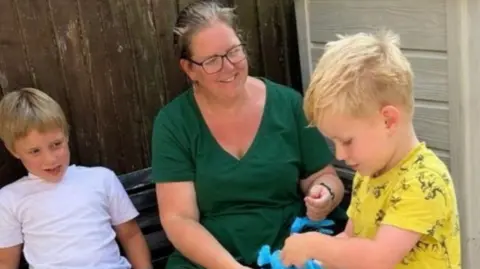 Supplied A woman wearing green, looking at a child wearing a yellow top who is holding a blue toy. There is a child wearing a white t-shirt looking at them both to the left