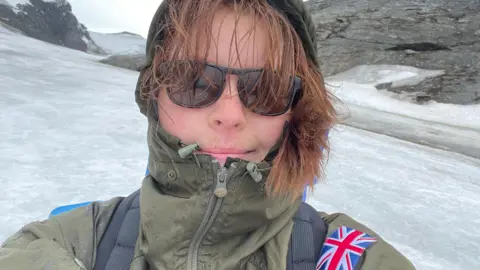 Emily Broadwell A selfie of Emily Broadwell wearing a khaki raincoat and black sunglasses with her hood up, and her gingery-brown hair poking out. Snow and ice on rocky mountains surround her.