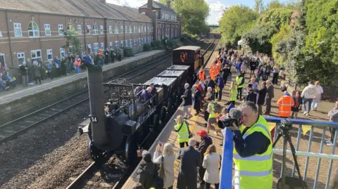 LDRS A historic steam locomotive stands at a railway station platform while large crowds gather on both sides of the tracks. Railway staff in high-visibility clothing and photographers are visible among spectators observing the heritage train.
