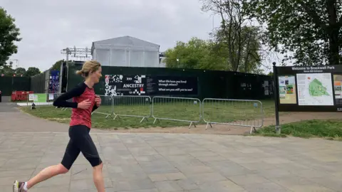 A photo taken of Brockwell Park during a previous festival season. A woman jogs next to barriers and a wall. A stage can be seen in the distance. 