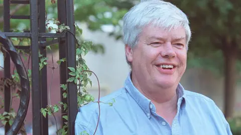 Michael Stuparyk A man with white hair smiles next to a black gate. He is wearing a light-blue shirt, and there are trees behind him.