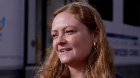 Katharine Field has long red hair and is looking to the right of the camera while being interviewed in front of the mobile chemotherapy unit.