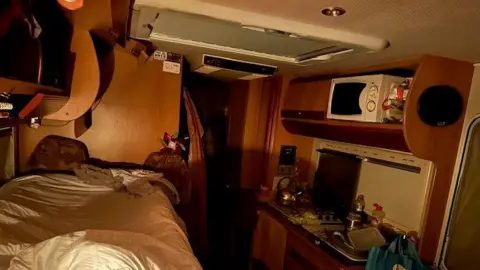 JO DUNNE The inside of the camper van. It it cramped and the is a bed opposite a small sing with a white microwave on a shelf above.