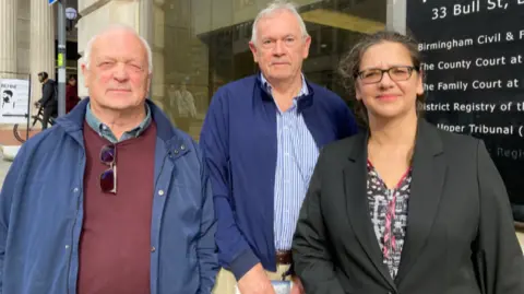 Amanda is wearing a grey jacket with a patterned top underneath and has glasses on. She has brown hair tied back. She is stood outside the High Court entrance with two men. Both wearing are blue jackets and shirts.