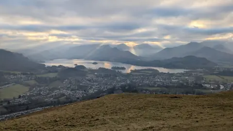 A view over Keswick taken from Latrigg Fell in the Lake District. The town sprawls from the foot of the fell, towards Derwentwater with more fells in the background. The sky is cloudy with rays of light shining through them.