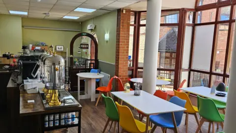 The inside of a cafe, showing tables chairs, with a counter, a brown door open, a large window and green walls. The flood is wood effect. 