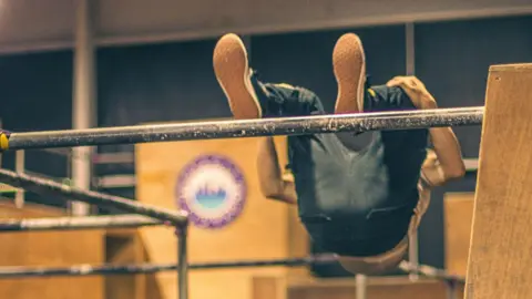 Lisa Beebe A man flips over a metal bar in jeans and trainers surrounded by wooden blocks