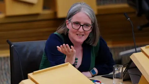 PA Media Maree Todd, a woman wearing glasses and with greying hair, sitting at a desk in the Scottish Parliament. She is speaking and has a hand out, gesturing with her palm open.