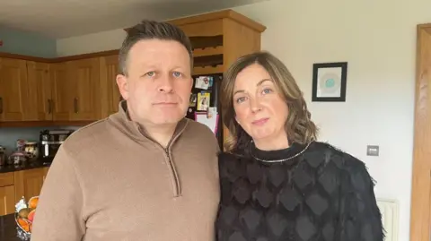 BBC A couple standing in a kitchen looking into the camera. The man has a brown jumper on with a zip. He has brown hair and blue eyes. He is pictured beside his wife. She has brown hair with blonde highlights, blue eyes and is wearing a silver necklace. There are kitchen cupboards in the background. 