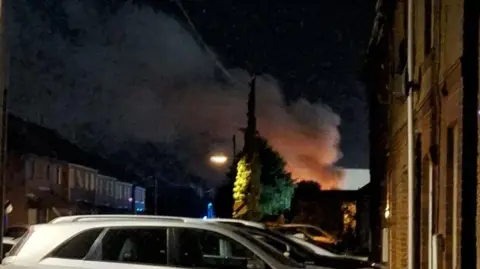 Sarah Worley The picture of a housing area taken at night-time. There are cars and vans parked on both sides of the street. In the background, a plume of orange smoke is billowing into the sky.