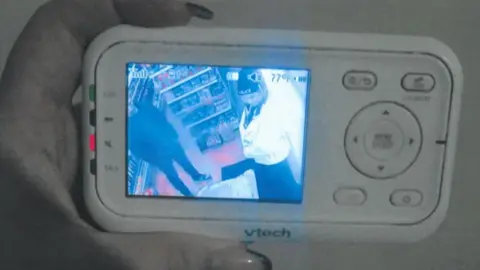 Trading Standards A handheld baby monitor displays a grainy security‑style video feed showing two people inside a shop.