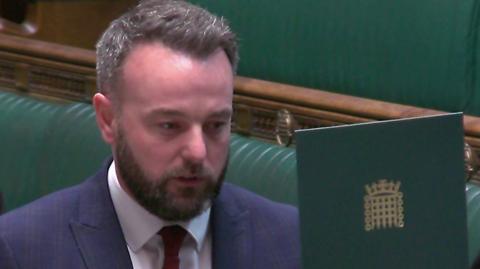 Colum Eastwood - a man with dark hair wearing a navy suit and red tie reads from a green cardboard sign with a gold embellishment of the House of Commons logo