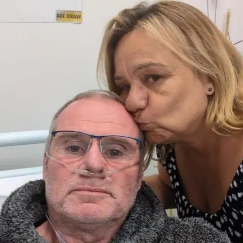 Matthew and Nicola Smith A man with a breathing tube in his nose looks at the camera while his wife, who has shoulder-length blonde hair, kisses him on the forehead. He is sat on a hospital bed.