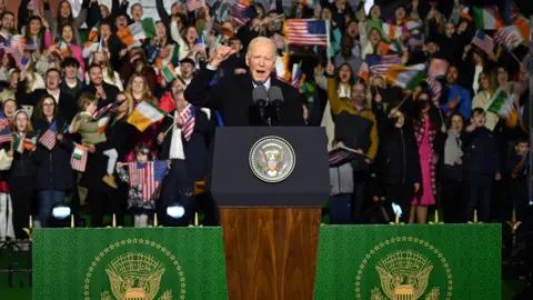 Leon Neal/Getty Images Joe Biden stands at a lecture addressing a large crown of well-wishers in Ballina, County Mayo, in 2023. He had short, white hair and is wearing a long black coat over a shirt and tie. People in the crowd behind him are waving US and Irish flags.