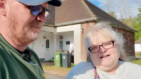A red-headed man wearing a cap and glasses with a beard looking off-camera, stands next to a woman with blond hair and glasses