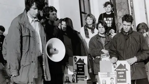 Chris Reynolds The black and white image shows a protest taking place on the steps of the then Welsh Office. On the left a campaigner is speaking into a loud-speaker. On the right a number of young people are listening on. They include Rhun ap Iorwerth who is holding a placard.