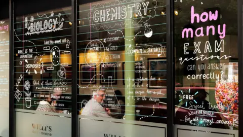 Gareth Williams Photography The window says Biology and Chemistry in white letters. People can be seen sitting in the cafe. Questions on the glass include what is the definition of osmosis and which of these decreases photosynthesis? There are drawings of a plant and chemistry bottles. 