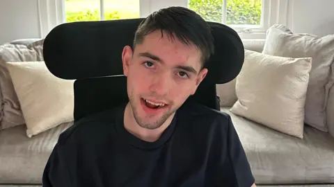 Lewis Syme sits in his chair, looking up to the camera - he is wearing a black t-shirt in a beige/cream living room - a sofa and cushions behind him.