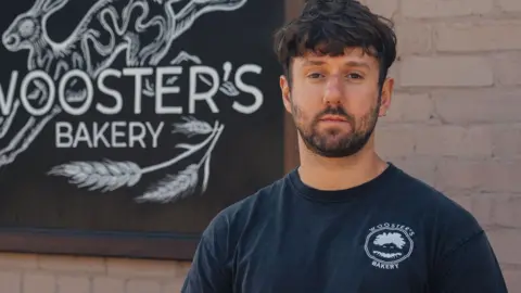 Supplied A head and shoulders image of a man wearing a black T-shirt while standing in front of a brick wall on which there is a sign that says Wooster's Bakery. 