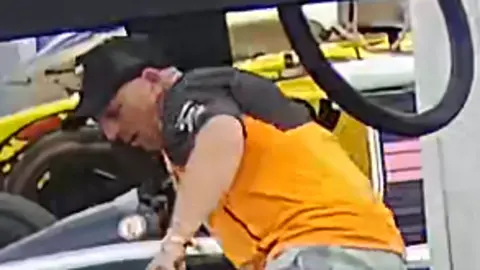 Northamptonshire Police A CCTV image of a man police wish to speak to after a classic F1 car was damaged at Silverstone on Friday. The man wears a black baseball cap and orange and black T-shirt while leaning over a car.