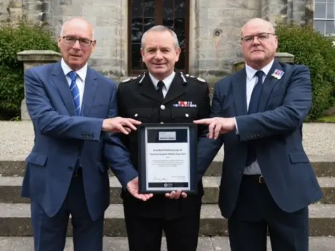 Adrian Hunt Two men wearing similar powder blue suits stand either side of the former Police Scotland Chief Constable Iain Livingstone, holding an award.