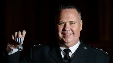 PA Media A man dressed in formal police uniform is smiling at the camera. He is holding up a king's police medal in his right hand. It's a small silver coin hung on a white and navy striped ribbon. The man has short, dark hair which is turning grey at the sides. He is stood against a black background. 