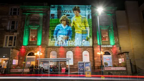 Night-time front view of the Filmhouse on Edinburgh's Lothian Road. The building is lit with red and green lights and an image from the film Gregory's Girl is projected on the outside. Its windows are boarded up.
