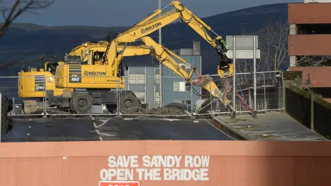 Large diggers ripping up a road. The road is blocked by a barrier on which someone has written "Save Sandy Row, open the bridge". There is a Road closed sign in front of that. In the background some hills.