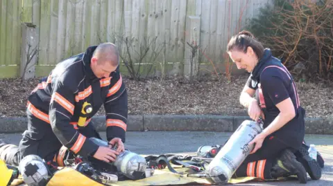 Cambridgeshire Fire and Rescue Service A man and a woman (Ms Bourne) are kneeling on the ground on a yellow mat. They are each holding a silver tank, about the size of a fire extinguisher, with Cambridgeshire Fire and Rescue Service written on. They are wearing blue fire service uniforms during the training exercise.