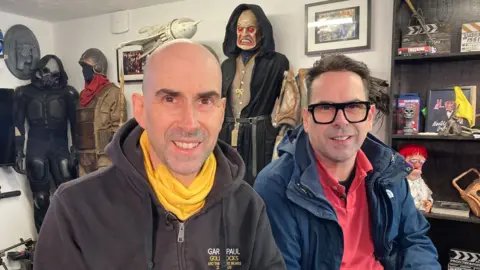 Paul and Gary sit in their office surrounded by science-fiction costumes, masks and props