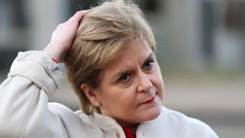 Getty Images A woman pushes short fair hair back on her head. She is wearing a cream jacket
