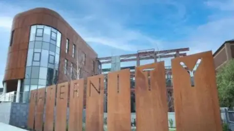 Belfast Trust The maternity hospital at the Royal Victoria Hospital. A multi-story building with copper coloured cladding and large windows. In the foreground there is grey pavement and a copper coloured sign spelling out the word maternity. 