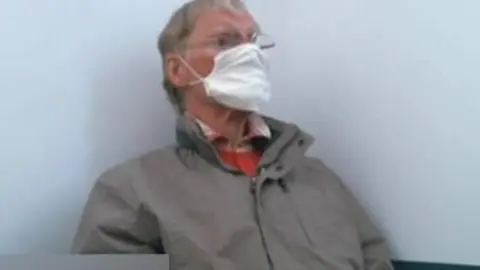 An old man wearing a face mask and a grey jacket sits against a white wall. He is in a police interview room. 