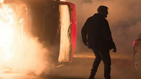 The figure of a character standing in silhouette on a road at night in front of a burning coach and a crashed car, in a scene from Corriedale