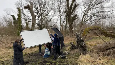 Harry Tomlin A wild Norfolk landscape including woodland, fallen trees leading to grassland. In the middle of the image is a film crew with a large light reflector filming a scene for the film. 