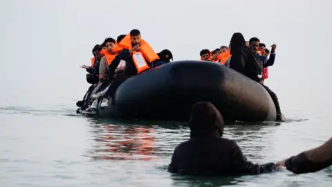 An inflatable taxi-boat filled with people approaches a person standing with their back to the camera, with water up their chest